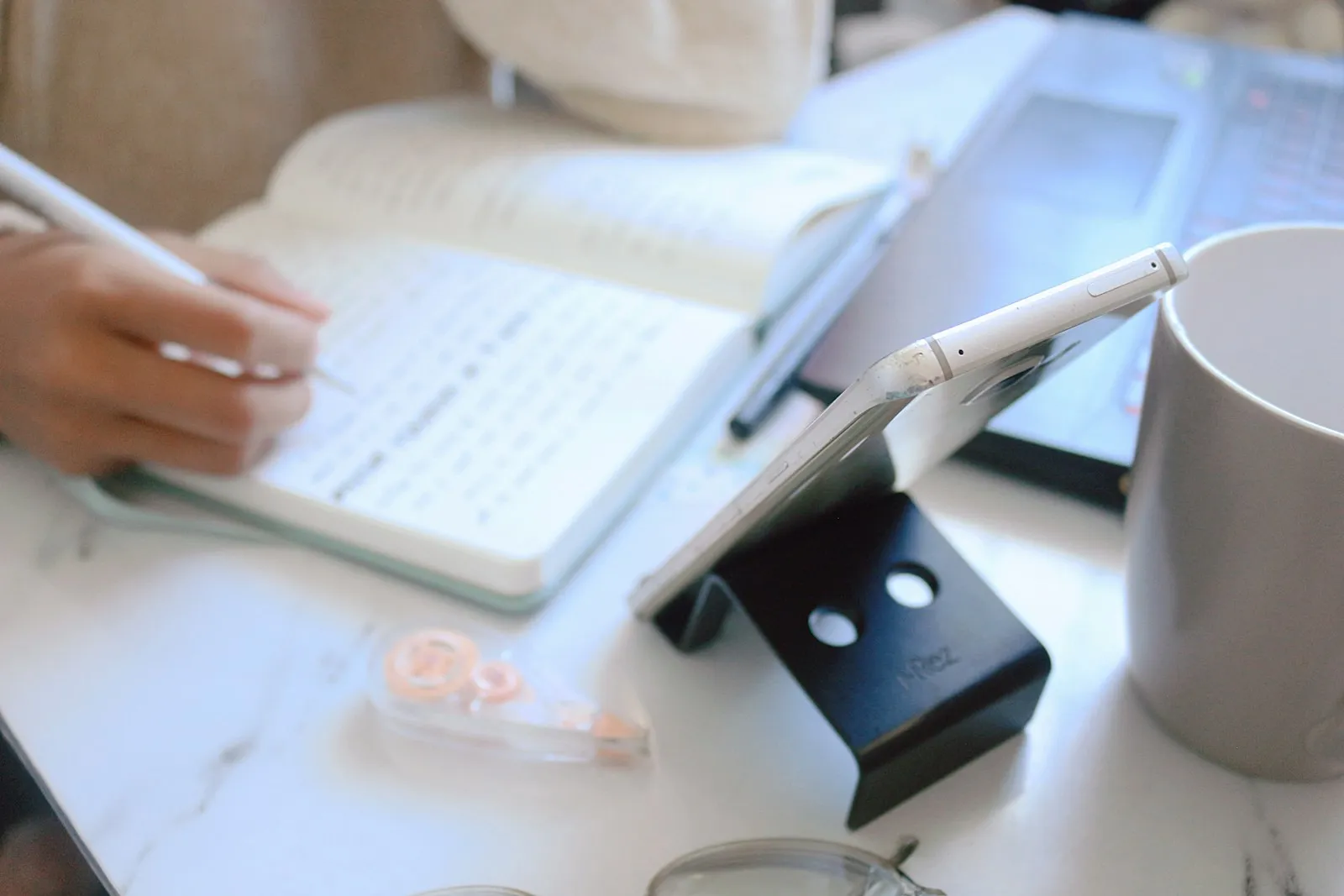 Una mano escribiendo notas junto a un computador y un teléfono